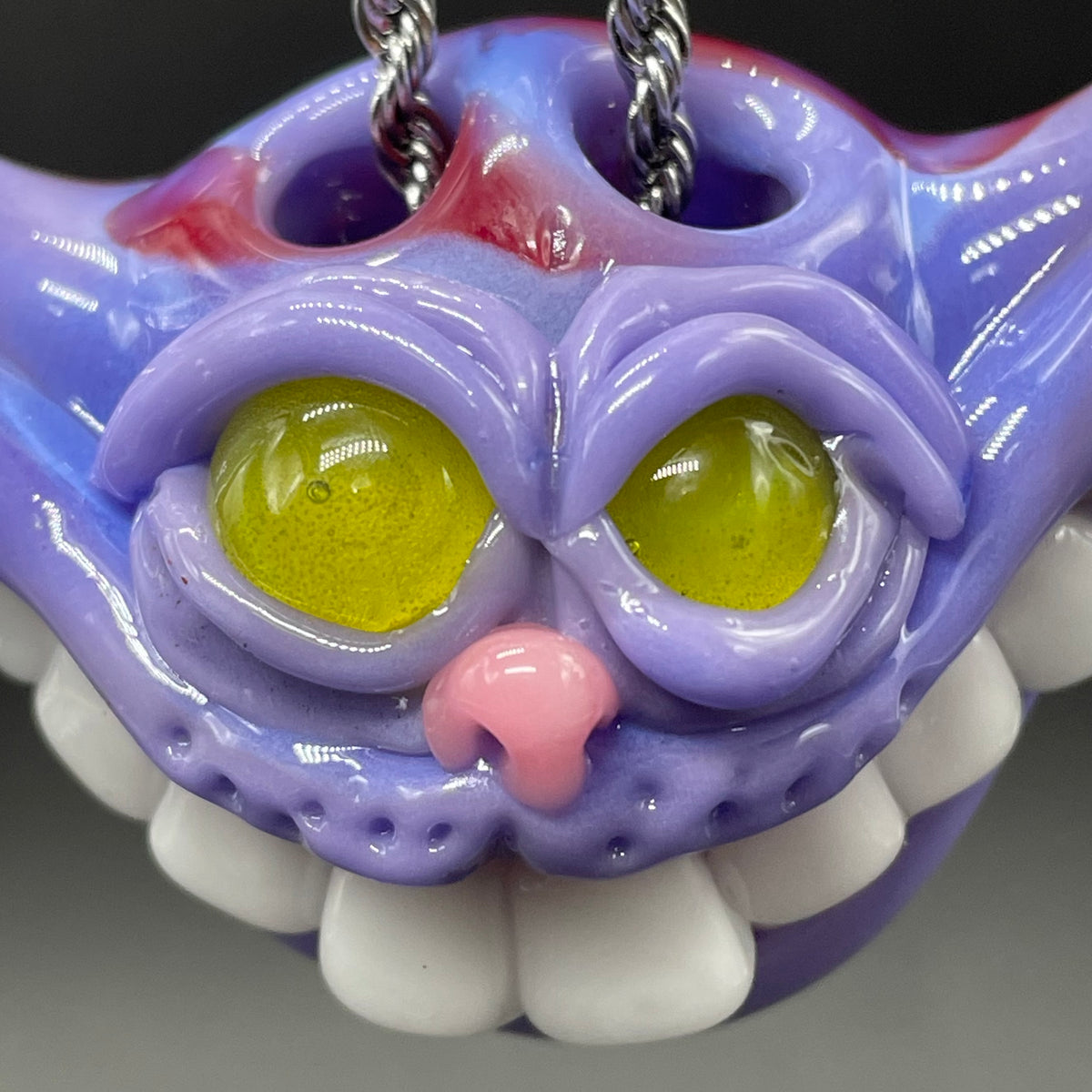 A close-up of a colorful, glossy monster skull keychain. The skull features a purple base with yellow-green eyes, a pink nose, white teeth, and red and purple shading on the forehead, with a silver chain visible at the top.