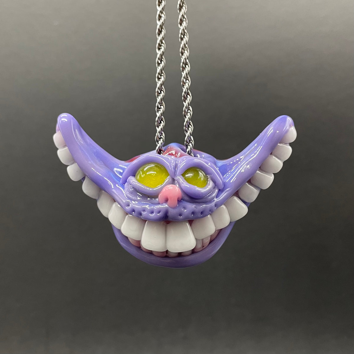 A purple Cheshire Cat mouth pendant necklace with white teeth, yellow eyes, and a silver chain, shown against a dark gray background.
