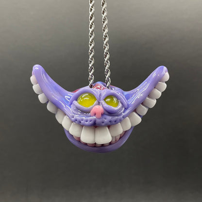 A purple Cheshire Cat mouth pendant necklace with white teeth, yellow eyes, and a silver chain, shown against a dark gray background.
