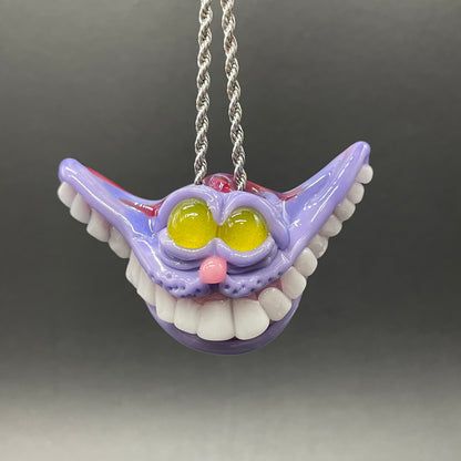 A hand blown glass pendant featuring a Cheshire Cat with a mischievous exaggerated wide, grinning cat with white realistic teeth, a small pink nose, and two large, glossy yellow eyes set in a lavender-colored cat  face. The pendant is suspended from a silver-toned twisted metal chain necklace.