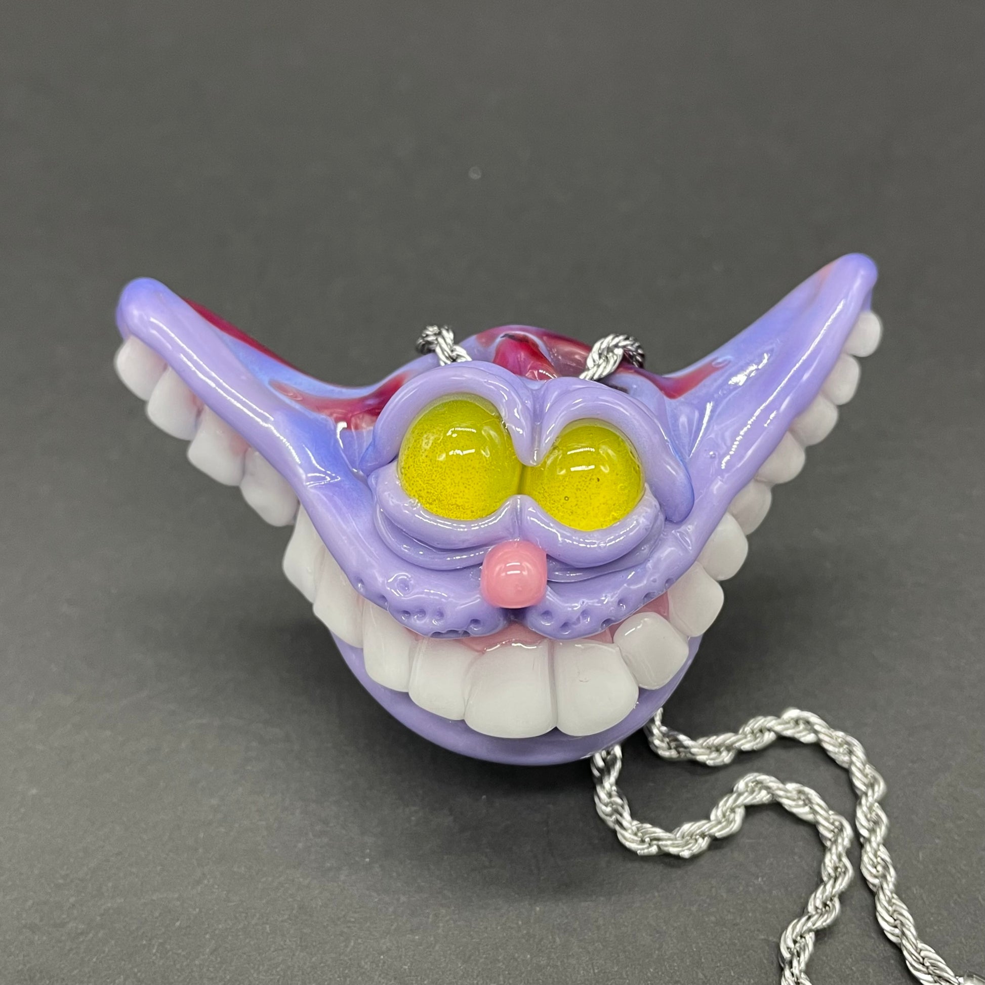 A hand blown glass pendant featuring a Cheshire Cat with a mischievous exaggerated wide, grinning cat with white realistic teeth, a small pink nose, and two large, glossy yellow eyes set in a lavender-colored cat  face. The pendant is suspended from a silver-toned twisted metal chain necklace.