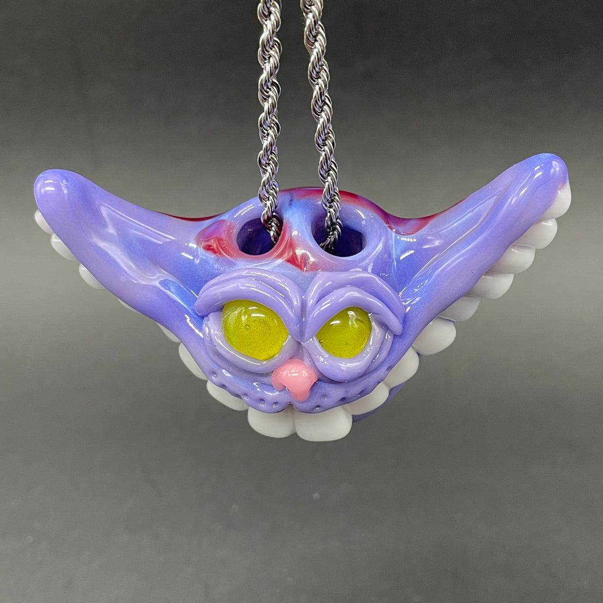 A whimsical cat-shaped glass pendant necklace on a silver chain, featuring a purple and lavender body with yellow eyes, a pink nose, white teeth, and red swirl details, set against a dark gray background.