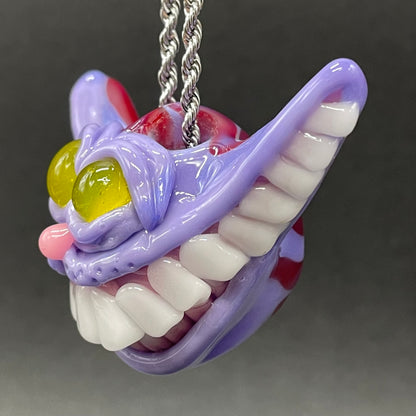 A hand blown glass pendant featuring a Cheshire Cat with a mischievous exaggerated wide, grinning cat with white realistic teeth, a small pink nose, and two large, glossy yellow eyes set in a lavender-colored cat  face. The pendant is suspended from a silver-toned twisted metal chain necklace.