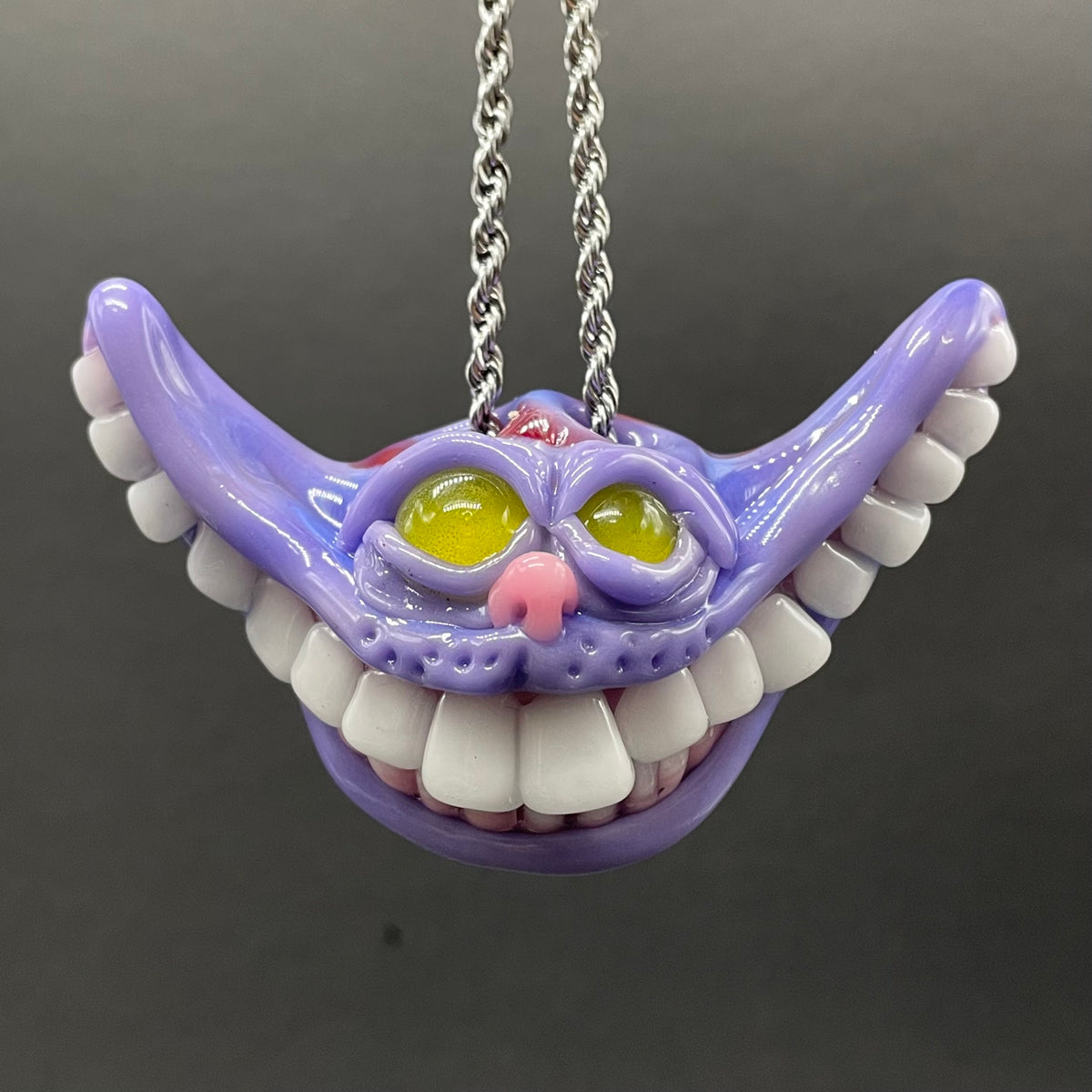 A purple Cheshire Cat style pendant necklace with white teeth, yellow eyes, and a pink nose, hanging from a silver chain against a dark gray background.