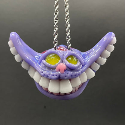 A purple Cheshire Cat style pendant necklace with white teeth, yellow eyes, and a pink nose, hanging from a silver chain against a dark gray background.