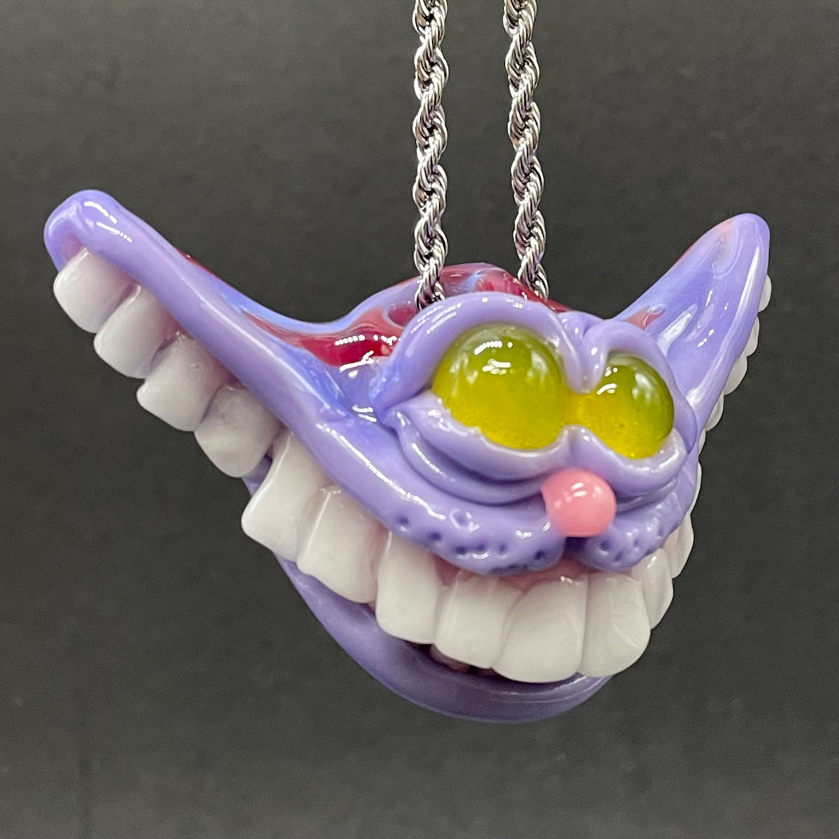 A hand blown glass pendant featuring a Cheshire Cat with a mischievous exaggerated wide, grinning cat with white realistic teeth, a small pink nose, and two large, glossy yellow eyes set in a lavender-colored cat  face. The pendant is suspended from a silver-toned twisted metal chain necklace.