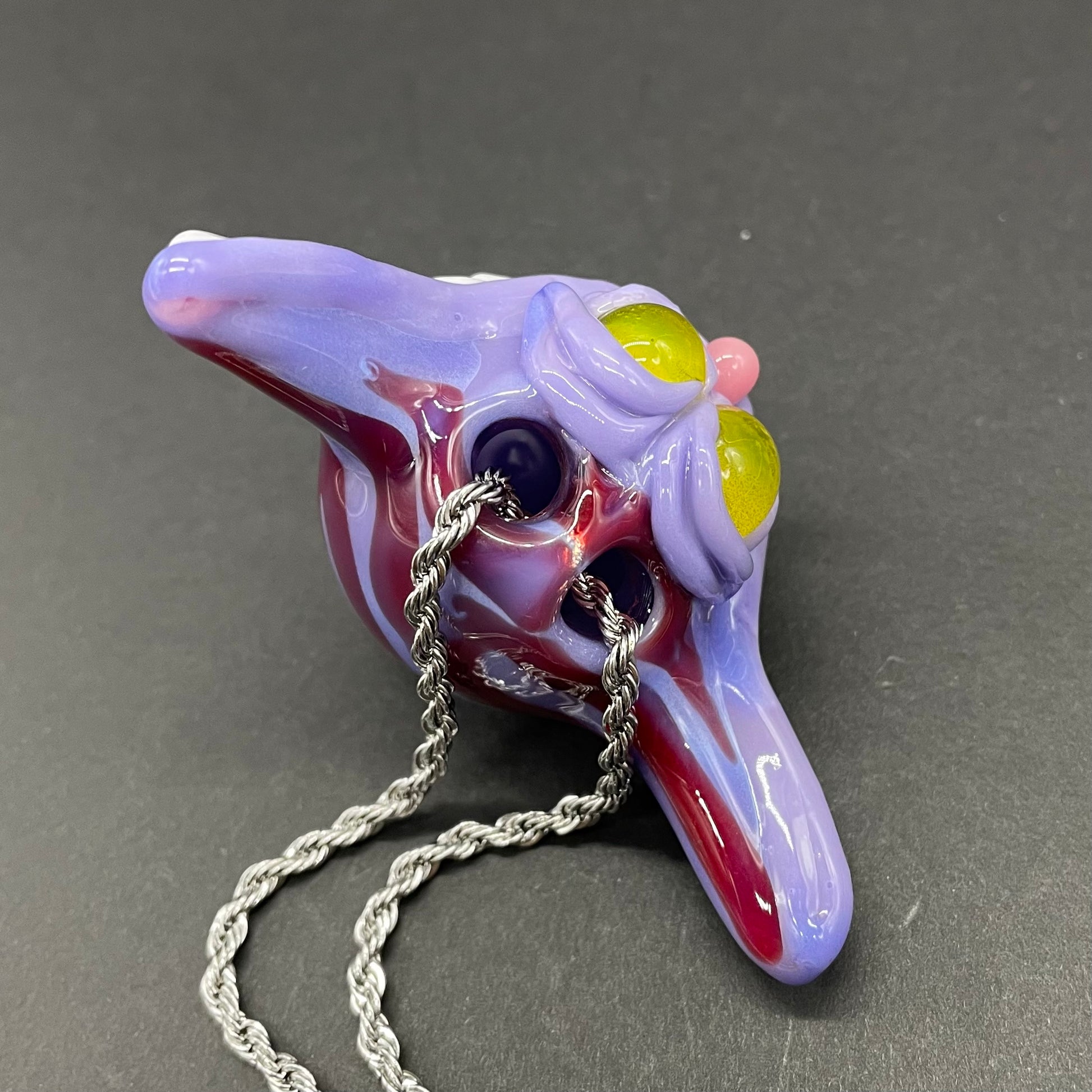 A hand blown glass pendant featuring a Cheshire Cat with a mischievous exaggerated wide, grinning cat with white realistic teeth, a small pink nose, and two large, glossy yellow eyes set in a lavender-colored cat  face. The pendant is suspended from a silver-toned twisted metal chain necklace.