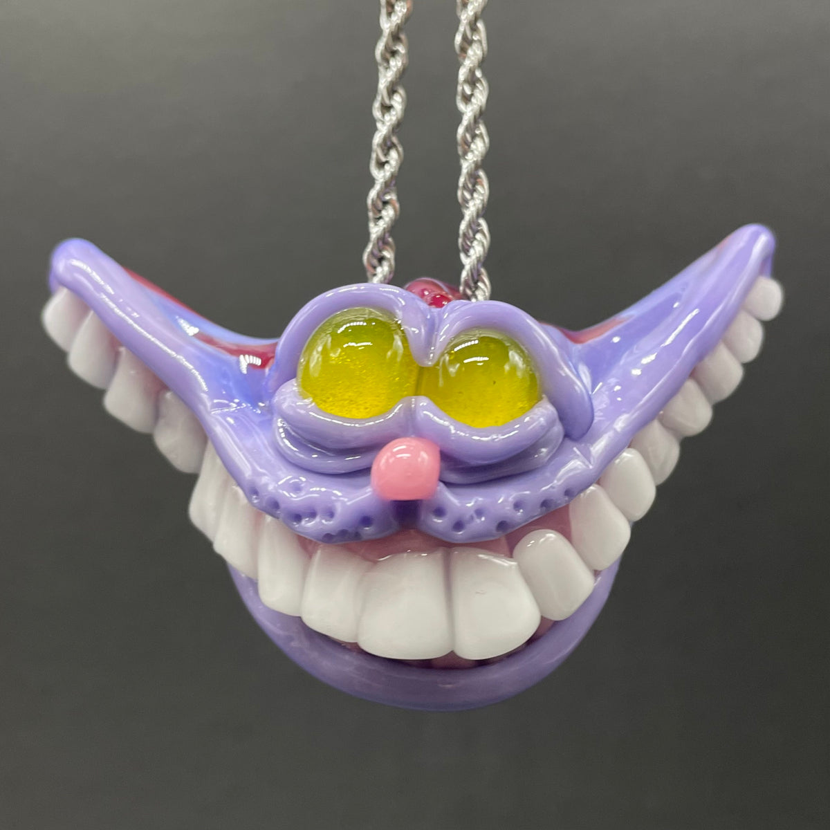 A hand blown glass pendant featuring a mischievous exaggerated wide, grinning cat with white realistic teeth, a small pink nose, and two large, glossy yellow eyes set in a lavender-colored cat  face. The pendant is suspended from a silver-toned twisted metal chain necklace.