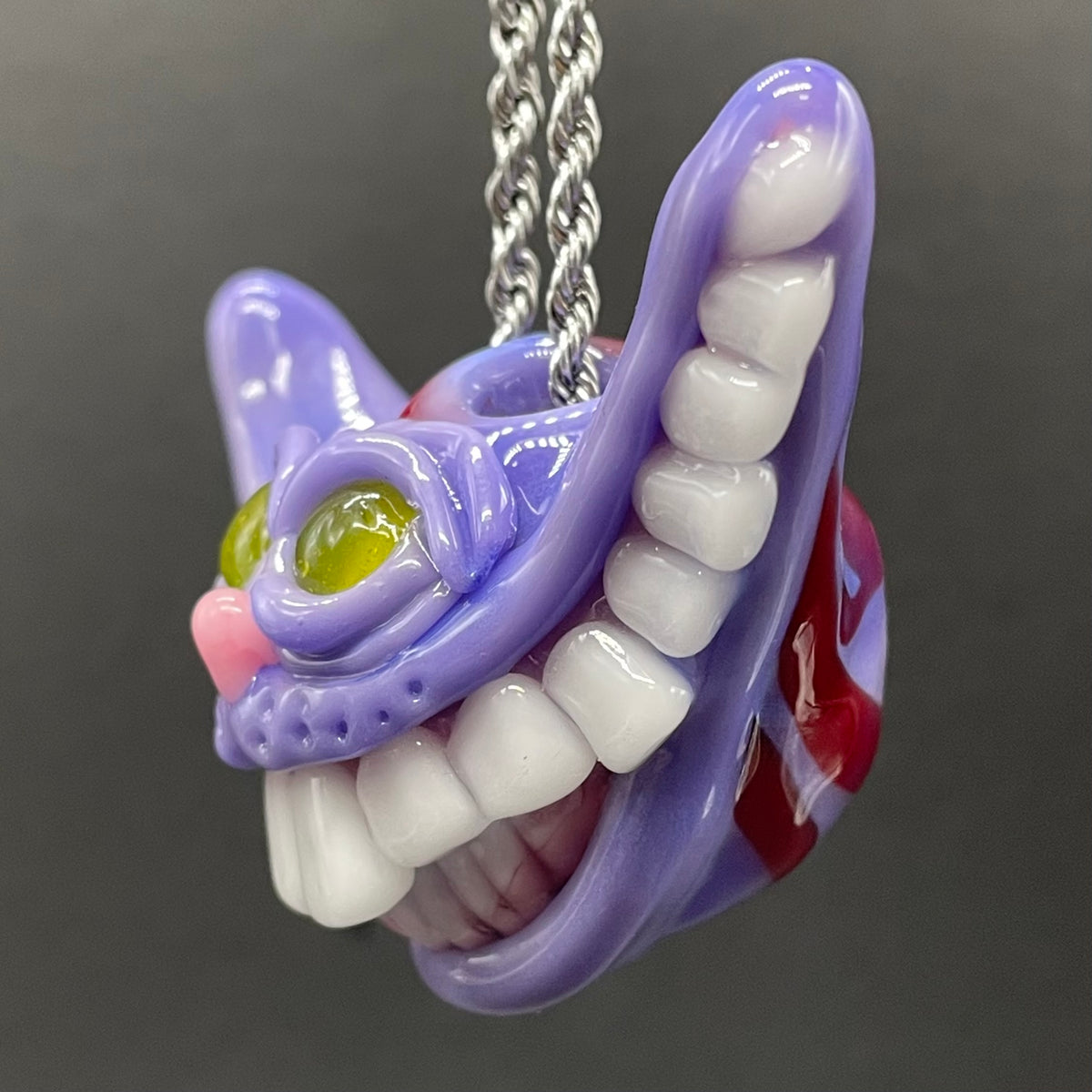 A close-up of a purple and white glass pendant shaped like an open mouth with large white teeth, a green eye, pink nose, and red inner details, hanging from a silver chain against a dark gray background.