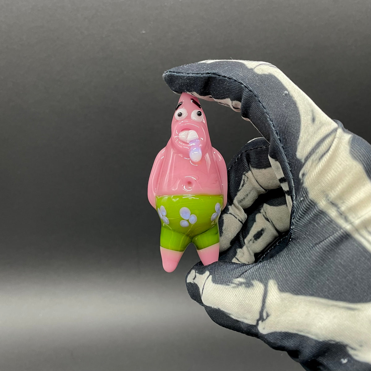 A small Patrick Star figurine held between a black and white tie-dye fabric glove against a dark gray background.