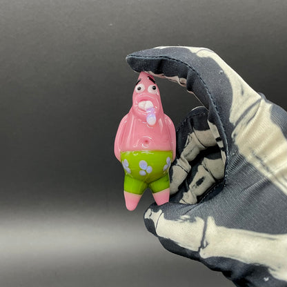 A small Patrick Star figurine held between a black and white tie-dye fabric glove against a dark gray background.