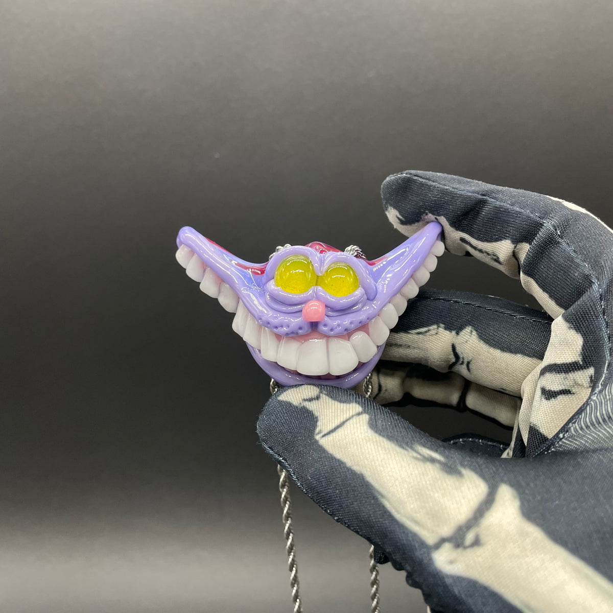 A hand blown glass pendant featuring a Cheshire Cat with a mischievous exaggerated wide, grinning cat with white realistic teeth, a small pink nose, and two large, glossy yellow eyes set in a lavender-colored cat  face. The pendant is suspended from a silver-toned twisted metal chain necklace.