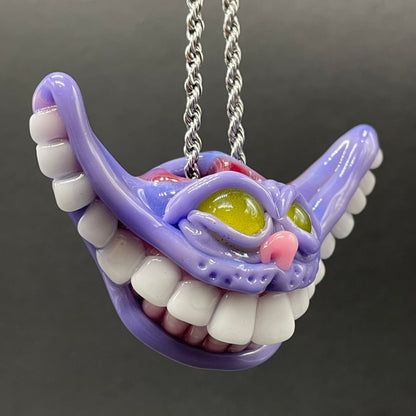 A purple Cheshire Cat-themed pendant necklace with white teeth, yellow-green eyes, and a pink nose, hanging from a silver chain against a dark gray background.