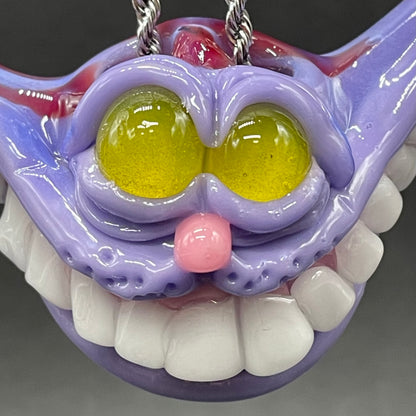 A hand blown glass pendant featuring a Cheshire Cat with a mischievous exaggerated wide, grinning cat with white realistic teeth, a small pink nose, and two large, glossy yellow eyes set in a lavender-colored cat  face. The pendant is suspended from a silver-toned twisted metal chain necklace.
