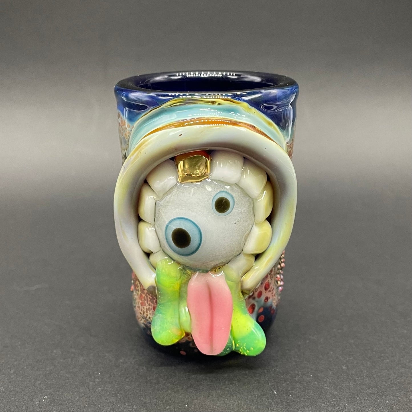 Blown glass cyclops monster hand blown shot glass with glow in the dark eyes inside a mouth with gold teeth. Green slimy drool flows out of its mouth with a long pink tongue. 