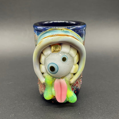 Blown glass cyclops monster hand blown shot glass with glow in the dark eyes inside a mouth with gold teeth. Green slimy drool flows out of its mouth with a long pink tongue. 