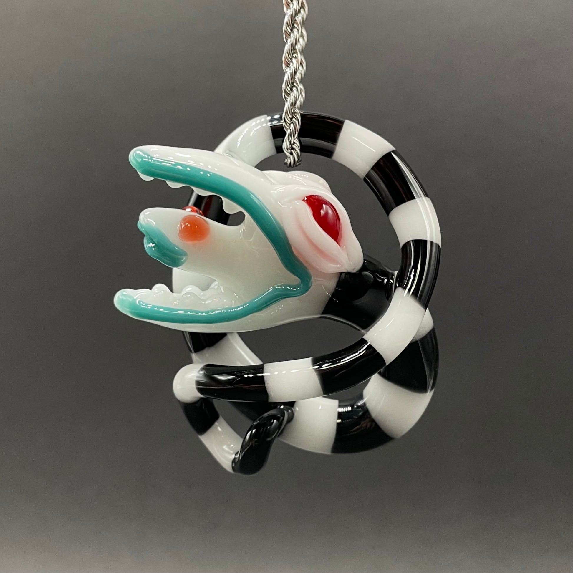 Beetlejuice Inspired handblown solid glass Sandworm sculpture - coiled black and white striped sandworm with white worm head, teal tongue, red eyes - Leviathan Glass

Beetlejuice sand worm hand blown glass pendant. contains black and white striped, twisting snake body. a large monster head with red eyes eating a smaller snake head in its mouth. 