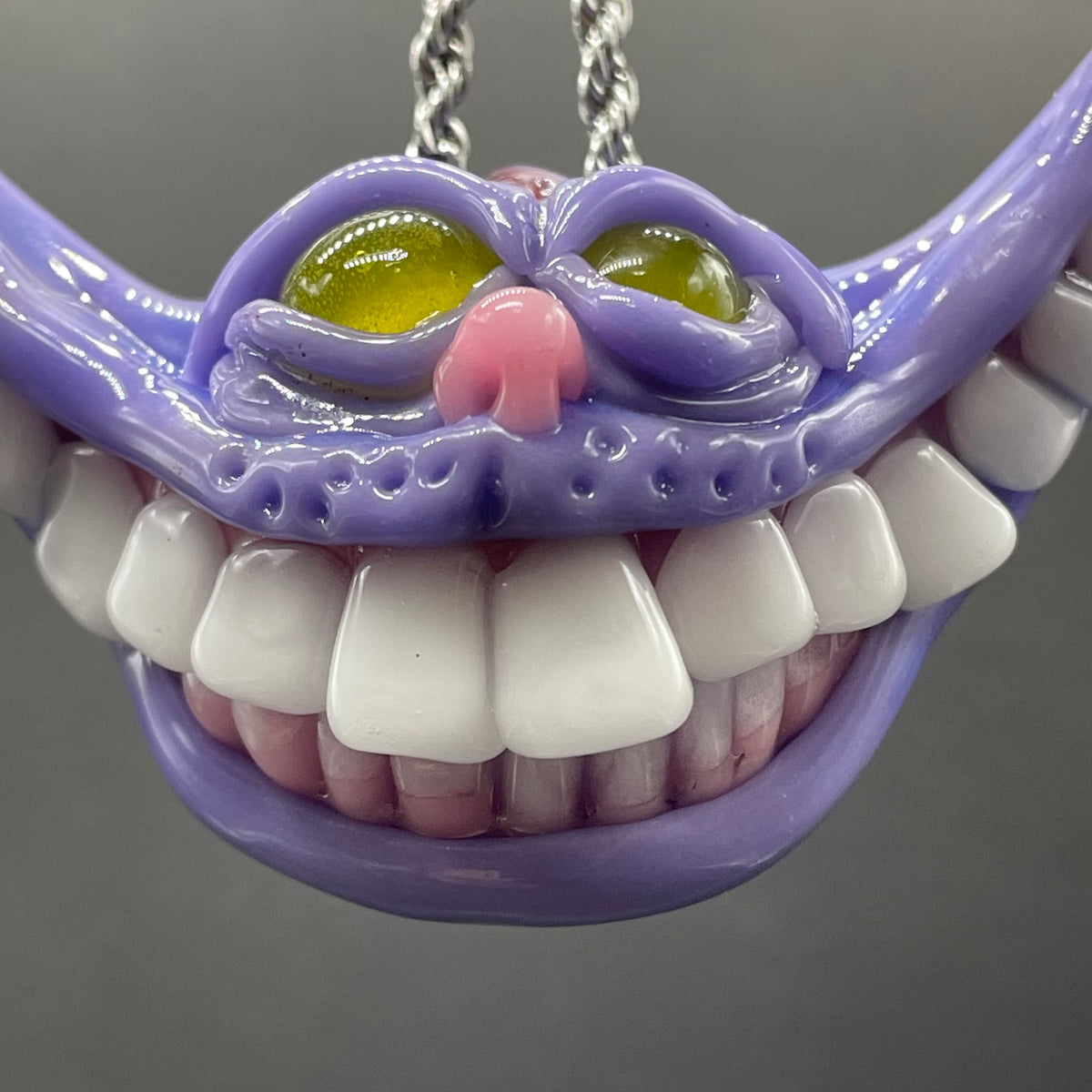 A close-up of a purple monster or alien mouth pendant with large white teeth, yellow-green eyes, and a pink nose, hanging from a silver chain against a gray background.