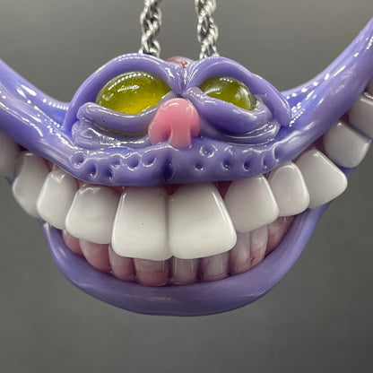 A close-up of a purple monster or alien mouth pendant with large white teeth, yellow-green eyes, and a pink nose, hanging from a silver chain against a gray background.