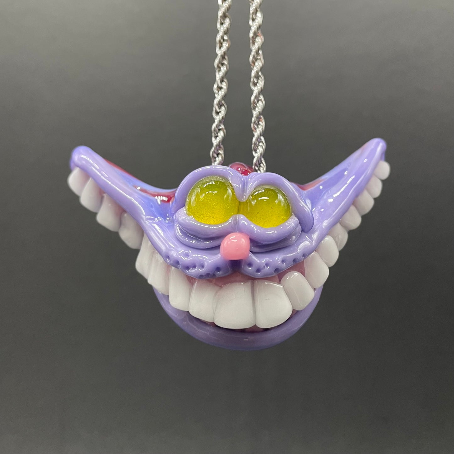 A hand blown glass pendant featuring a mischievous exaggerated wide, grinning cat with white realistic teeth, a small pink nose, and two large, glossy yellow eyes set in a lavender-colored cat  face. The pendant is suspended from a silver-toned twisted metal chain necklace.