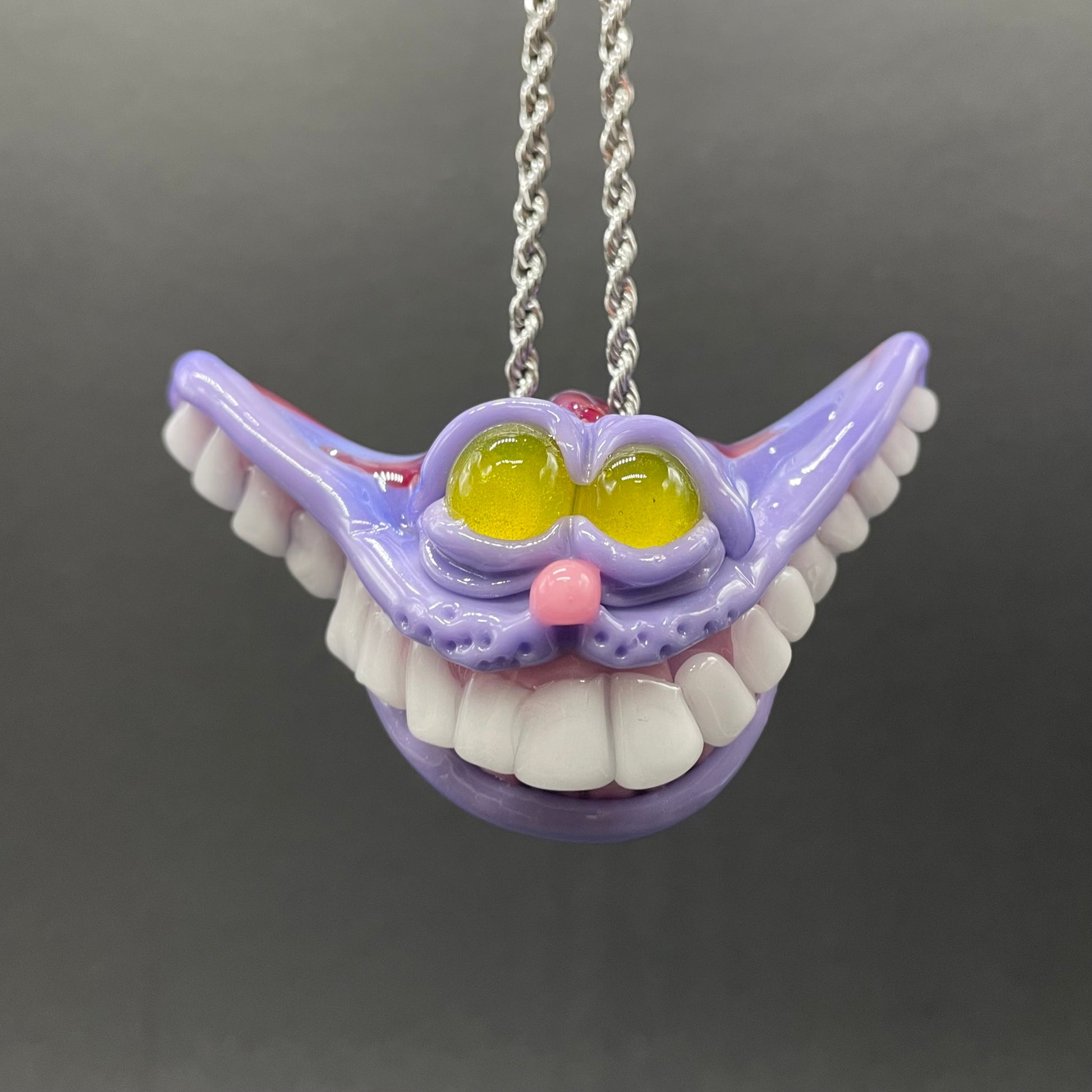 A hand blown glass pendant featuring a mischievous exaggerated wide, grinning cat with white realistic teeth, a small pink nose, and two large, glossy yellow eyes set in a lavender-colored cat  face. The pendant is suspended from a silver-toned twisted metal chain necklace.