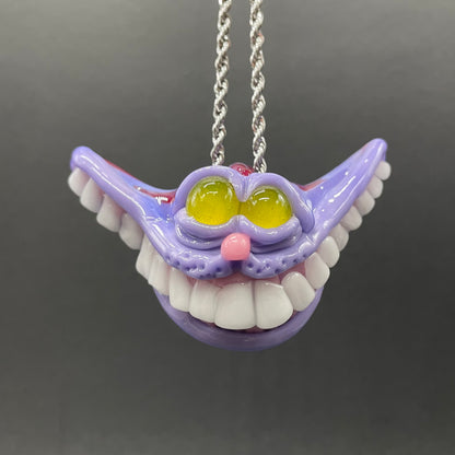 A hand blown glass pendant featuring a mischievous exaggerated wide, grinning cat with white realistic teeth, a small pink nose, and two large, glossy yellow eyes set in a lavender-colored cat  face. The pendant is suspended from a silver-toned twisted metal chain necklace.