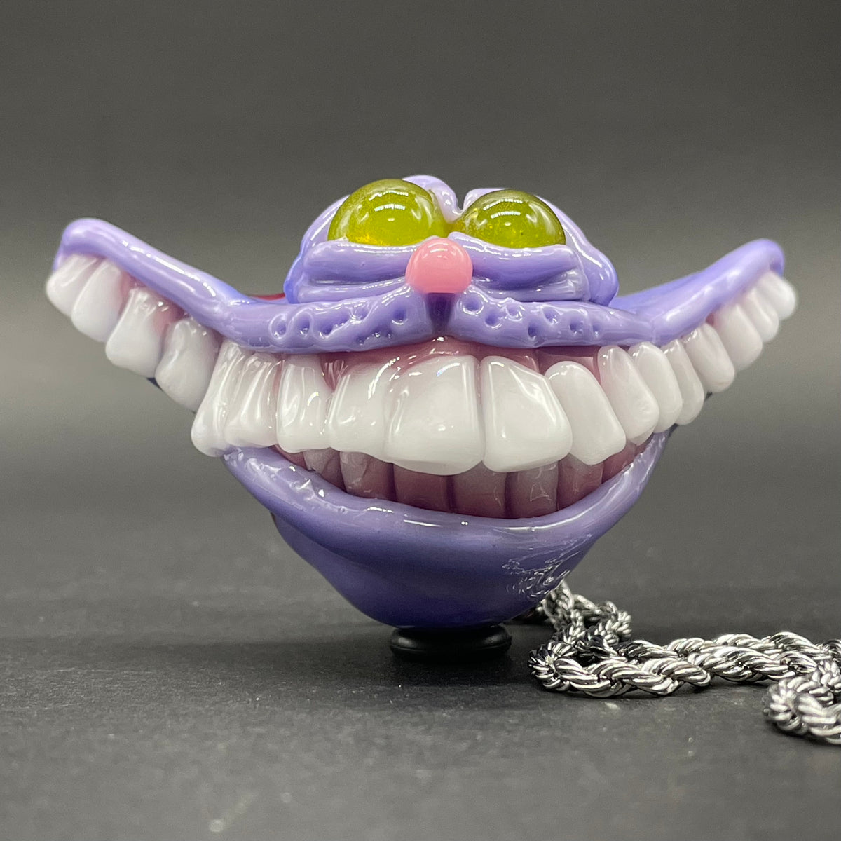 A hand blown glass pendant featuring a Cheshire Cat with a mischievous exaggerated wide, grinning cat with white realistic teeth, a small pink nose, and two large, glossy yellow eyes set in a lavender-colored cat  face. The pendant is suspended from a silver-toned twisted metal chain necklace.