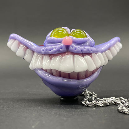 A hand blown glass pendant featuring a Cheshire Cat with a mischievous exaggerated wide, grinning cat with white realistic teeth, a small pink nose, and two large, glossy yellow eyes set in a lavender-colored cat  face. The pendant is suspended from a silver-toned twisted metal chain necklace.