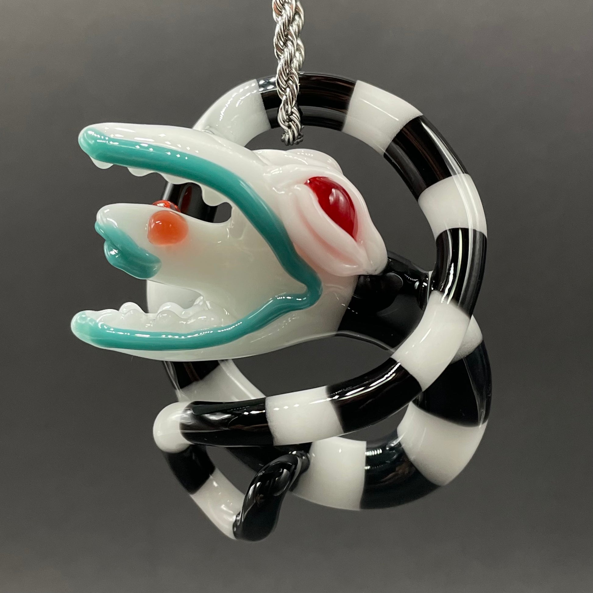 Beetlejuice Inspired handblown solid glass Sandworm sculpture - coiled black and white striped sandworm with white worm head, teal tongue, red eyes - Leviathan Glass
Beetlejuice sand worm hand blown glass pendant. contains black and white striped, twisting snake body. a large monster head with red eyes eating a smaller snake head in its mouth. 