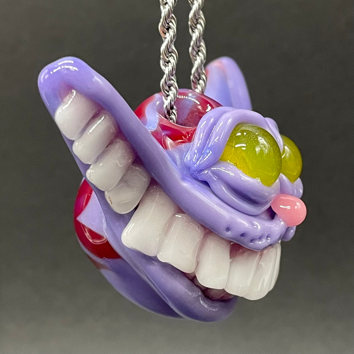 A hand blown glass pendant featuring a Cheshire Cat with a mischievous exaggerated wide, grinning cat with white realistic teeth, a small pink nose, and two large, glossy yellow eyes set in a lavender-colored cat  face. The pendant is suspended from a silver-toned twisted metal chain necklace.