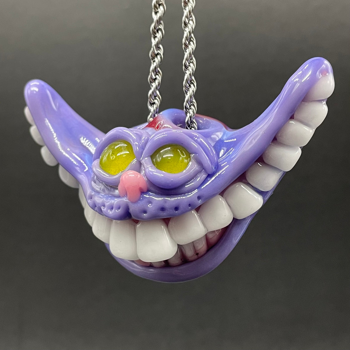 A purple Cheshire Cat style pendant necklace with white teeth, yellow eyes, and a pink nose, hanging from a silver chain against a dark gray background.