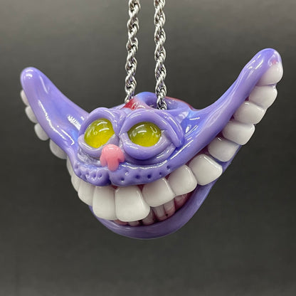 A purple Cheshire Cat style pendant necklace with white teeth, yellow eyes, and a pink nose, hanging from a silver chain against a dark gray background.