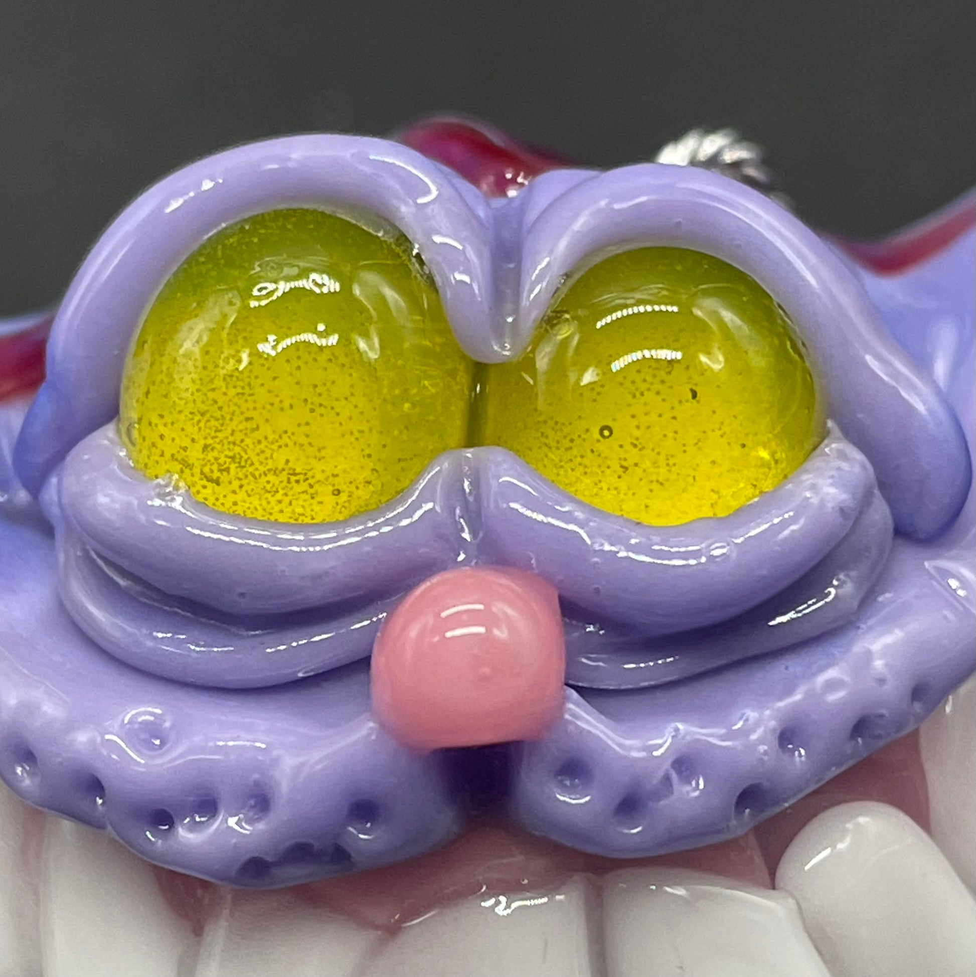 A hand blown glass pendant featuring a Cheshire Cat with a mischievous exaggerated wide, grinning cat with white realistic teeth, a small pink nose, and two large, glossy yellow eyes set in a lavender-colored cat  face. The pendant is suspended from a silver-toned twisted metal chain necklace.