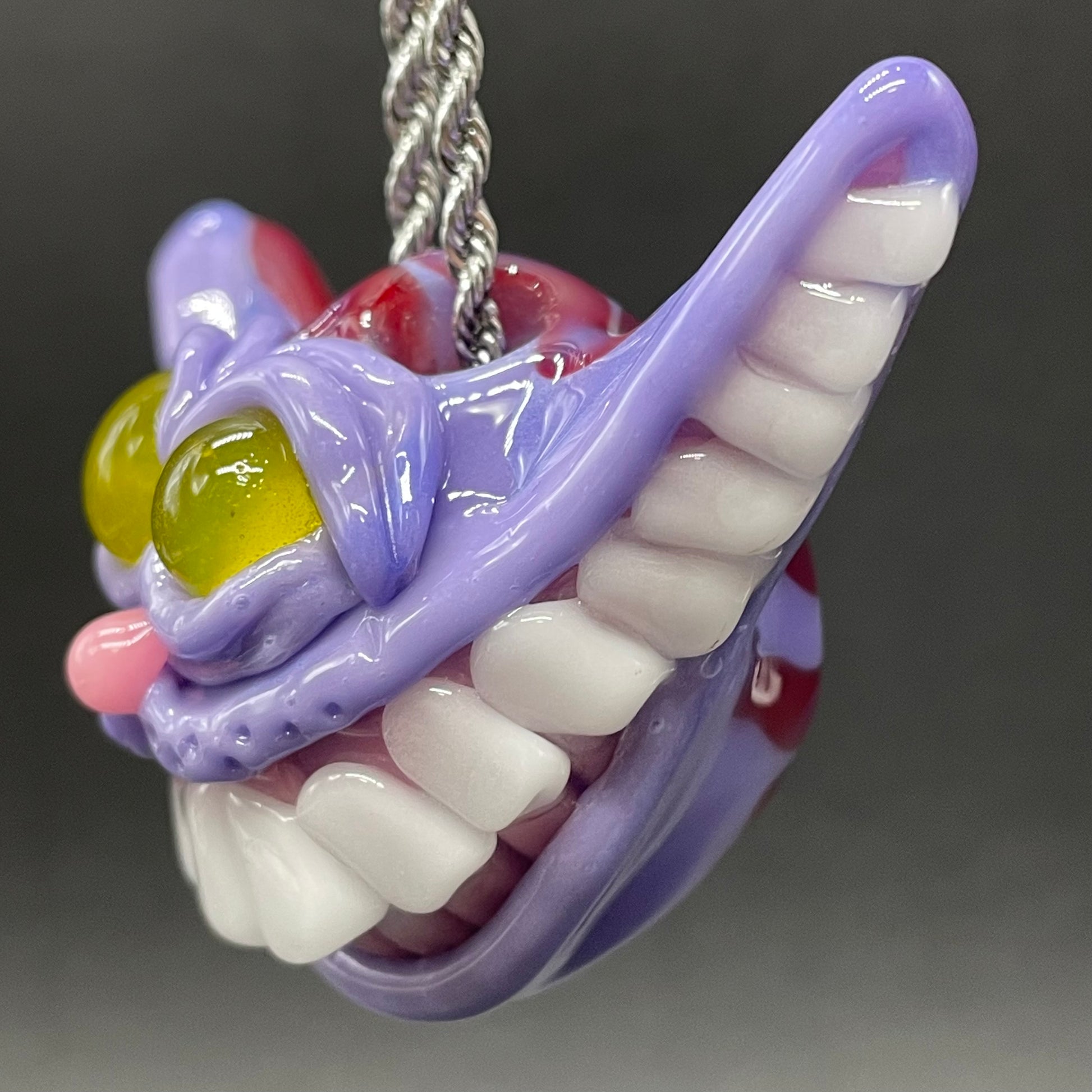 A hand blown glass pendant featuring a Cheshire Cat with a mischievous exaggerated wide, grinning cat with white realistic teeth, a small pink nose, and two large, glossy yellow eyes set in a lavender-colored cat  face. The pendant is suspended from a silver-toned twisted metal chain necklace.