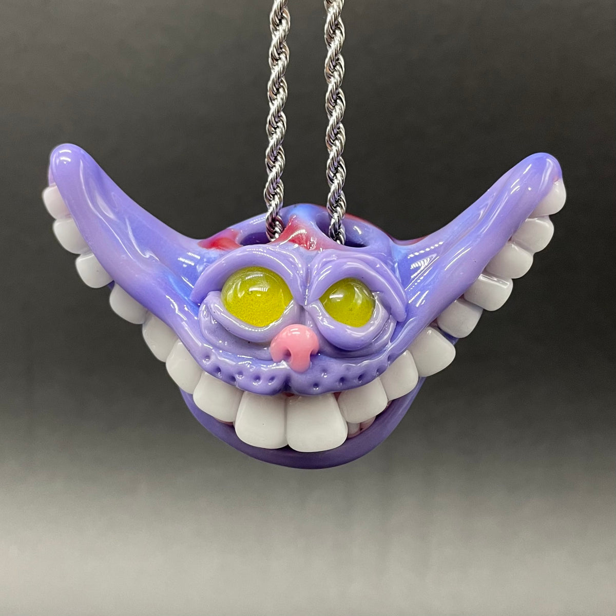A purple Cheshire Cat style pendant necklace with a silver twisted chain, featuring yellow eyes, a pink nose, and white teeth, shown against a dark gray gradient background.