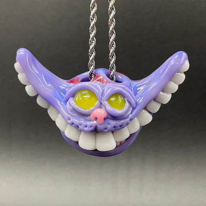 A purple Cheshire Cat style pendant necklace with a silver twisted chain, featuring yellow eyes, a pink nose, and white teeth, shown against a dark gray gradient background.