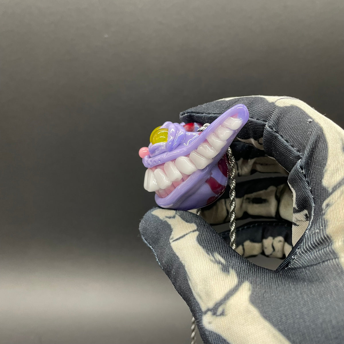 A hand blown glass pendant featuring a Cheshire Cat with a mischievous exaggerated wide, grinning cat with white realistic teeth, a small pink nose, and two large, glossy yellow eyes set in a lavender-colored cat  face. The pendant is suspended from a silver-toned twisted metal chain necklace.