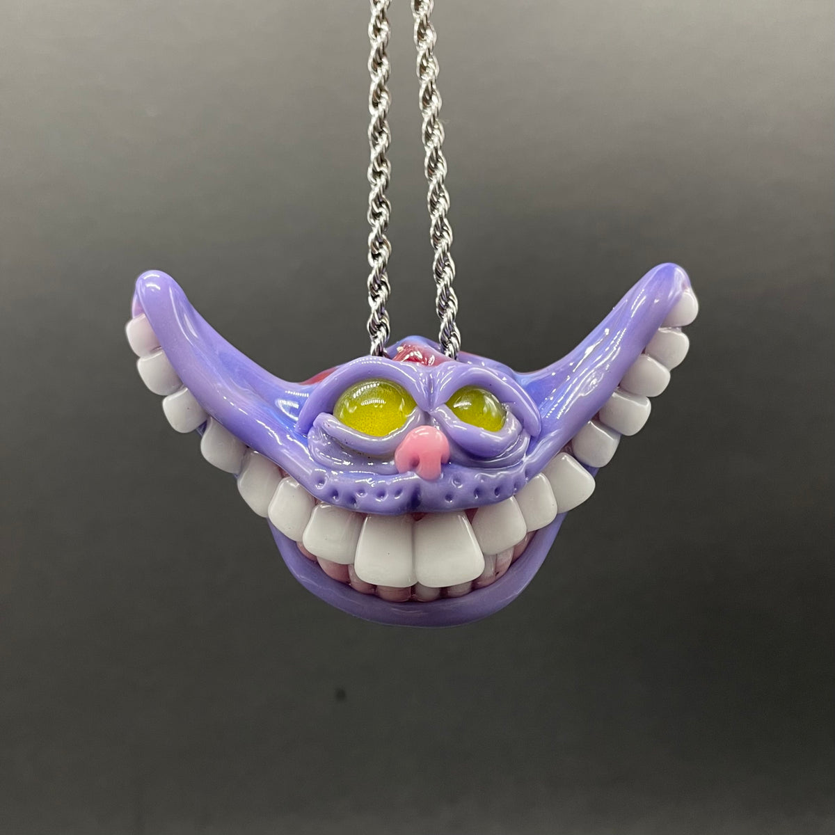 A purple Cheshire Cat-style toothy mouth pendant necklace with white teeth, yellow eyes, and a pink nose, hanging from a silver chain against a dark gray background.