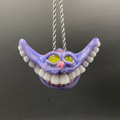 A purple Cheshire Cat-style toothy mouth pendant necklace with white teeth, yellow eyes, and a pink nose, hanging from a silver chain against a dark gray background.