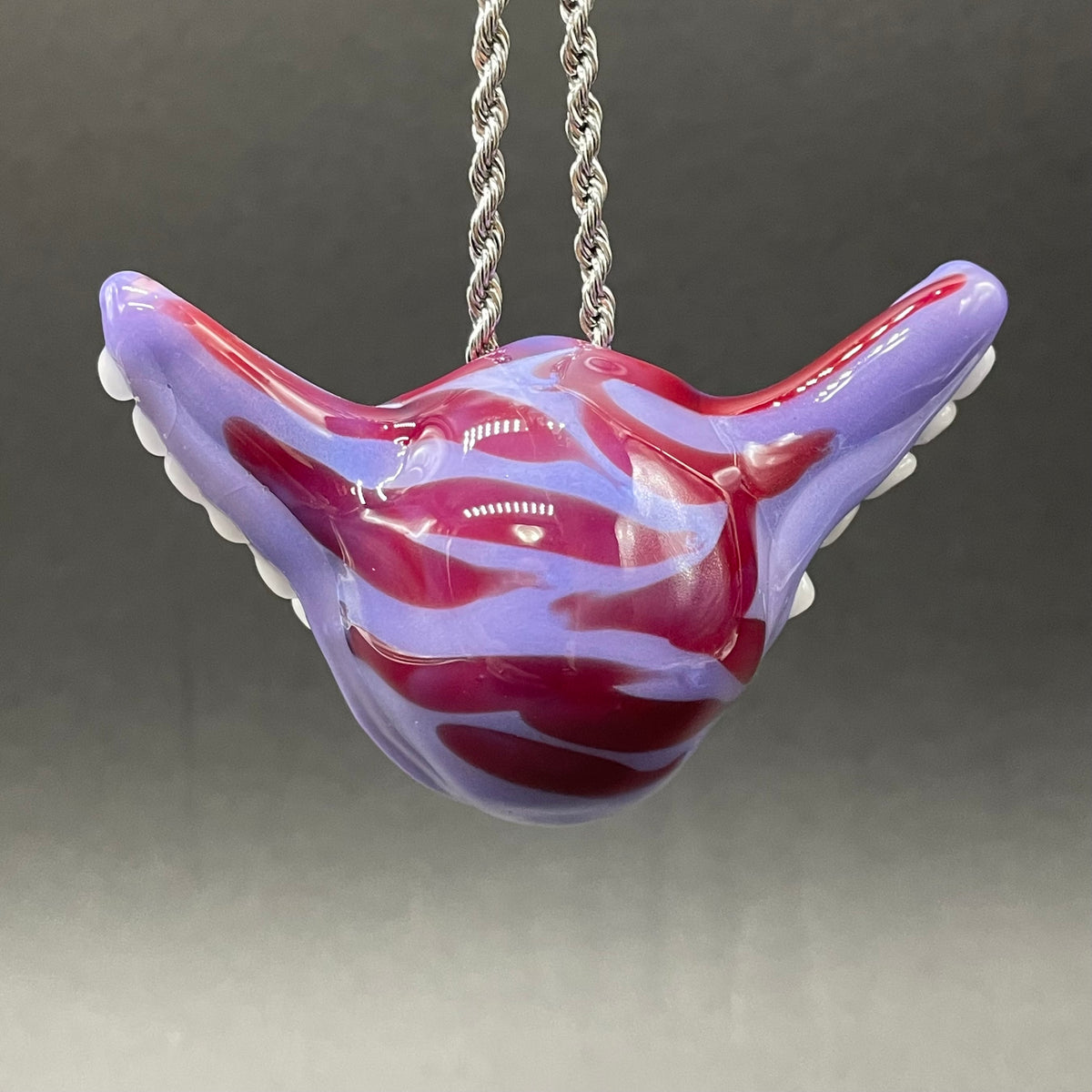 A hand blown glass pendant featuring a Cheshire Cat with a mischievous exaggerated wide, grinning cat with white realistic teeth, a small pink nose, and two large, glossy yellow eyes set in a lavender-colored cat  face. The pendant is suspended from a silver-toned twisted metal chain necklace.