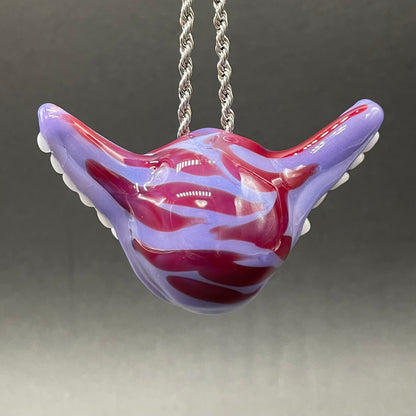 A hand blown glass pendant featuring a Cheshire Cat with a mischievous exaggerated wide, grinning cat with white realistic teeth, a small pink nose, and two large, glossy yellow eyes set in a lavender-colored cat  face. The pendant is suspended from a silver-toned twisted metal chain necklace.