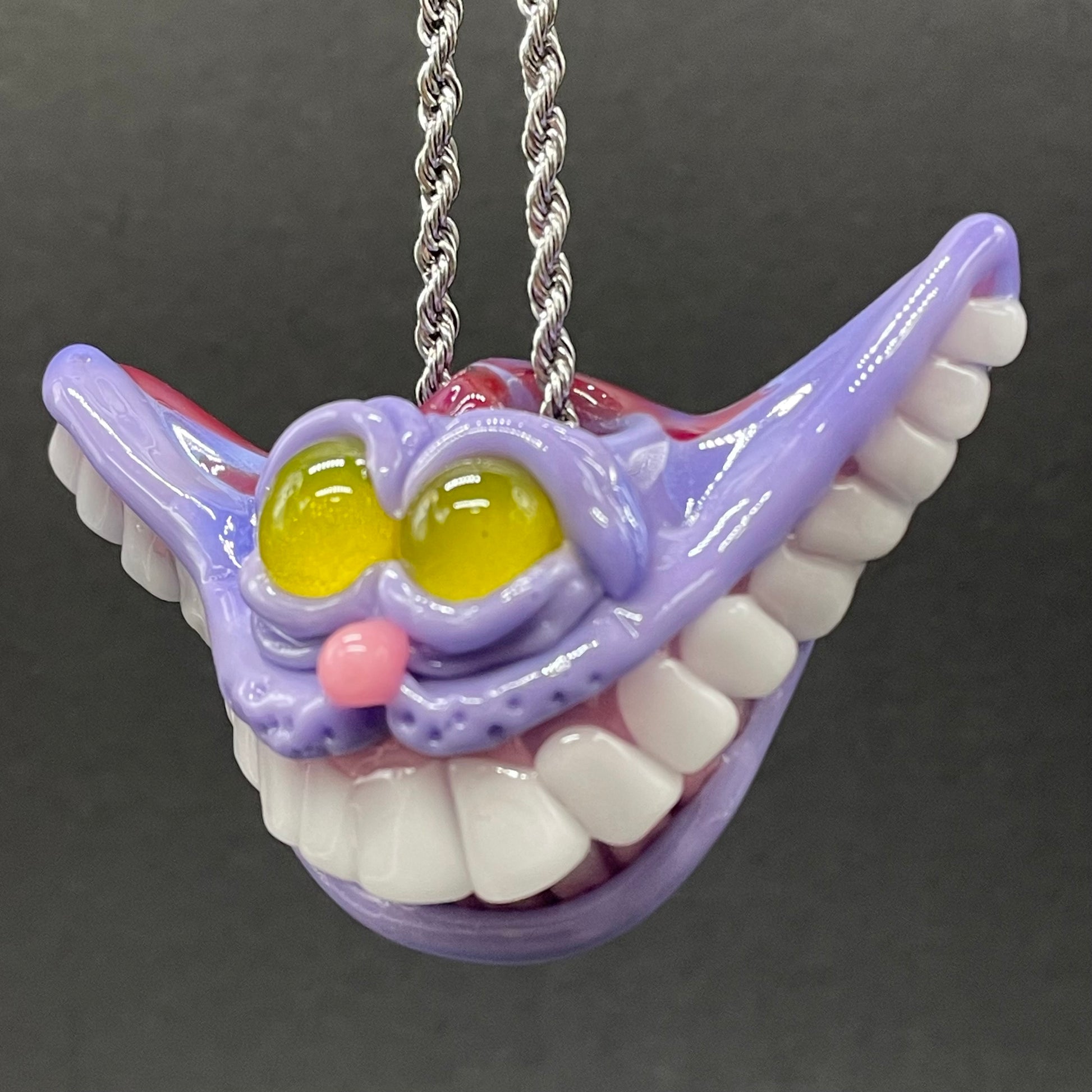 A hand blown glass pendant featuring a Cheshire Cat with a mischievous exaggerated wide, grinning cat with white realistic teeth, a small pink nose, and two large, glossy yellow eyes set in a lavender-colored cat  face. The pendant is suspended from a silver-toned twisted metal chain necklace.