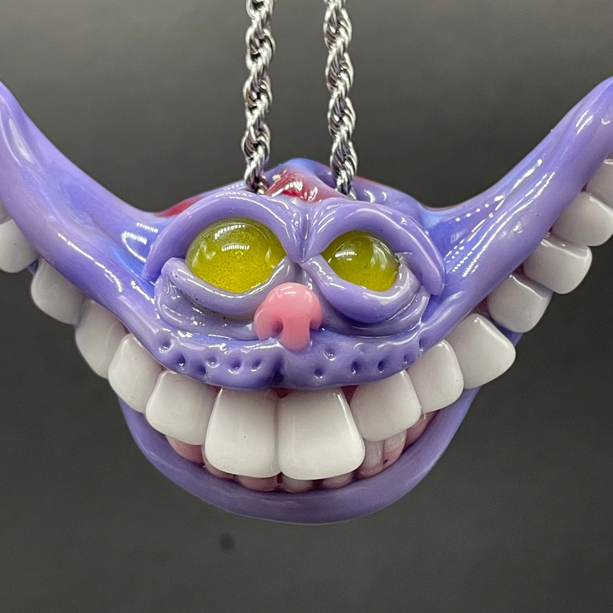 A purple Cheshire Cat style pendant necklace with white teeth, yellow eyes, and a pink nose, hanging from a silver chain against a dark gray background.
