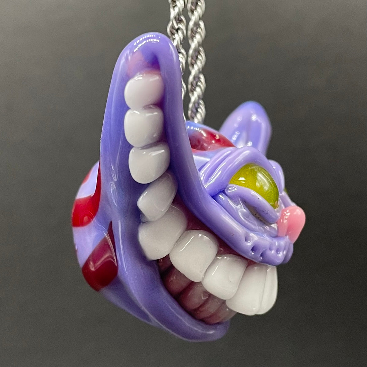 A close-up of a purple Cheshire Cat pendant necklace with white teeth, red and pink accents, and a green glass eye, hanging from a silver chain against a dark gray background.