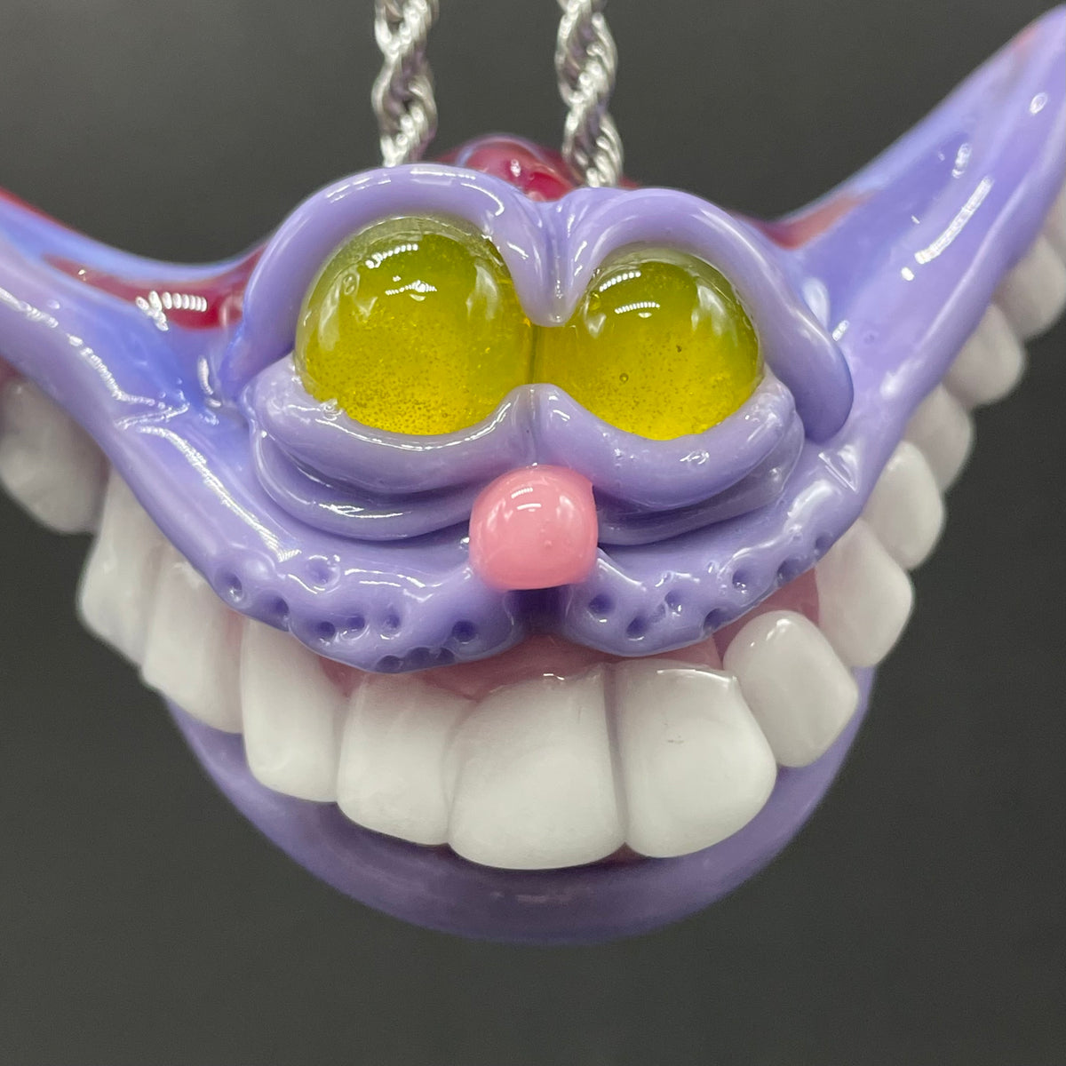 A hand blown glass pendant featuring a Cheshire Cat with a mischievous exaggerated wide, grinning cat with white realistic teeth, a small pink nose, and two large, glossy yellow eyes set in a lavender-colored cat  face. The pendant is suspended from a silver-toned twisted metal chain necklace.
