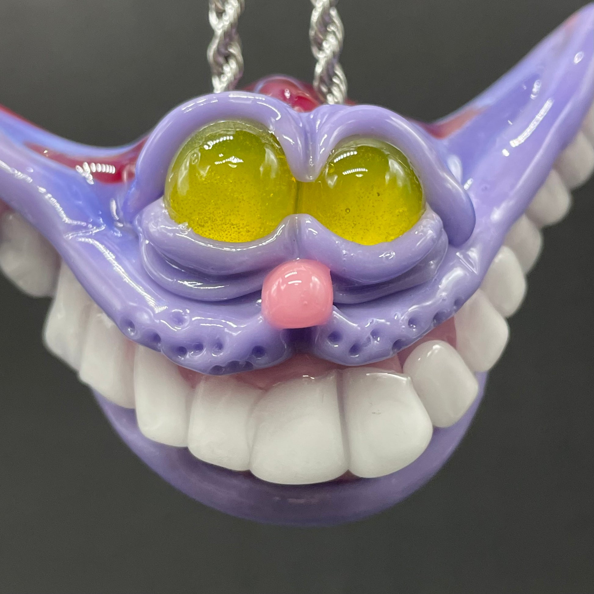 A hand blown glass pendant featuring a Cheshire Cat with a mischievous exaggerated wide, grinning cat with white realistic teeth, a small pink nose, and two large, glossy yellow eyes set in a lavender-colored cat  face. The pendant is suspended from a silver-toned twisted metal chain necklace.